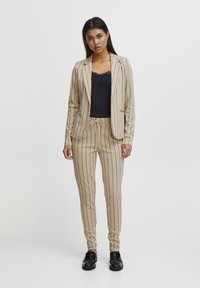 Beige gestreifter Anzug mit tailliertem Blazer und Hose mit Kordelzug, kombiniert mit einem schwarzen Spitzen-Camisole und schwarzen Schuhen. Glatte Textur durchgehend.