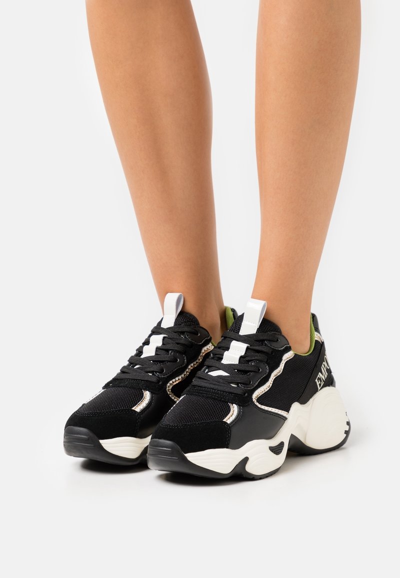 Svarta och vita klumpiga sneakers med snörningsdesign, bärs på bara ben mot en enkel vit bakgrund.