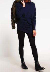 Marinblå oversized stickad tröja med rund halsringning, ihop med svarta skinny jeans och ankelstövlar. Olivgrön bomberjacka hängande över armen.