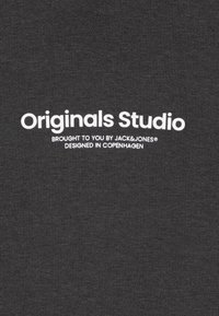 Tessuto grigio scuro con un knit testurizzato, caratterizzato da un testo bianco bold: "Originals Studio" e un testo più piccolo sottostante che indica il marchio e l'origine del design.