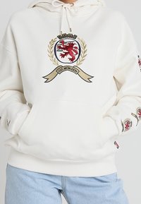 Felpa bianca con cappuccio caratterizzata da un ricamo con uno stemma raffigurante un leone rosso e accenti dorati. Include una tasca frontale e cappuccio con cordini.
