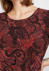 Czarna z czerwonym paisley wzorem bluzka z okrągłym dekoltem; gładki materiał; posiada skomplikowane kwiatowe i spirale wzory; krótkie rękawy.