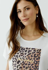 T-shirt blanc avec un graphique imprimé léopard et le texte "WILD THOUGHTS" en doré. Encolure ronde et texture de tissu douce.