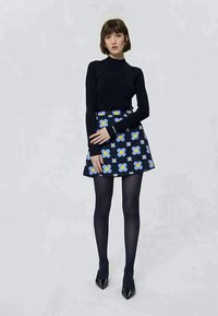 Mulher de pé, vestindo uma blusa preta de manga comprida, saia azul com padrão floral, collants pretos e sapatos pretos apontados, contra um fundo simples.