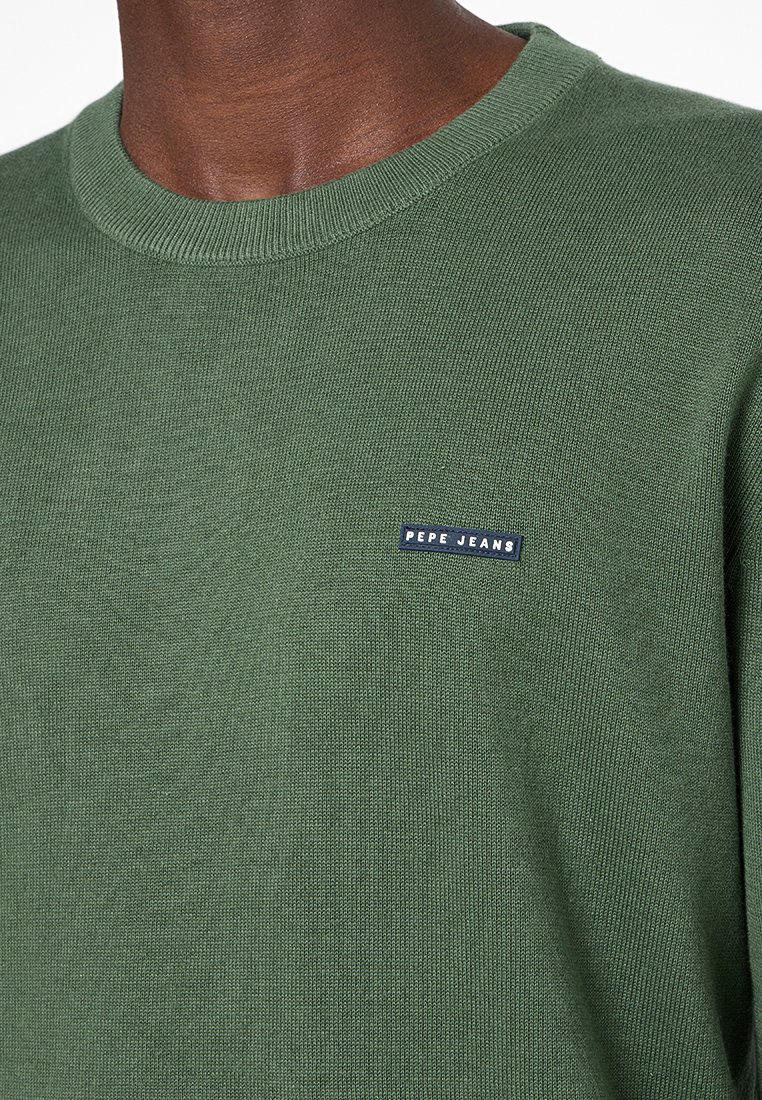 Pull en maille verte avec un col rond et des manches côtelées. Présente un patch rectangulaire bleu avec le texte "PEPE JEANS" sur la poitrine.