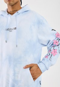 Sweatshirt azul com efeito tie-dye e bolso canguru, apresentando o texto preto "BERLIM" e bordado floral rosa ao longo da manga esquerda.