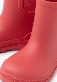 Czerwone gumowe buty z gładkim wykończeniem, z teksturowanymi akcentami i zaokrąglonym noskiem. Podeszwa ma wzorzystą powierzchnię dla lepszej przyczepności.