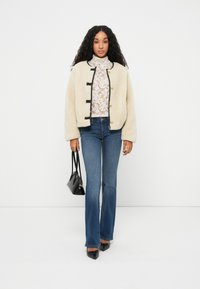 Cremefarvet faux fur jakke med sorte kanter og gyldne knapper, lagdelt over en hvid blonde top, parret med mørkeblå flagrende jeans og sorte hæle.