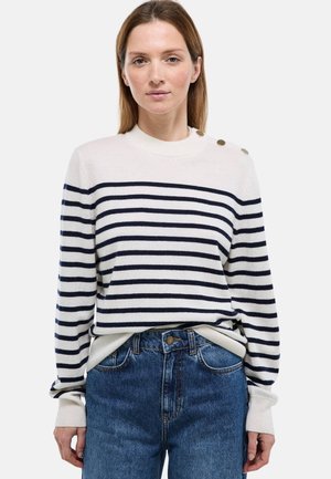 Femme portant un pull blanc à rayures bleu marine avec des boutons sur l'épaule et un jean bleu taille haute, debout devant un fond uni.