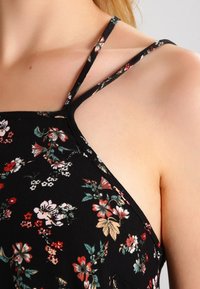 Gros plan sur une personne portant une robe noire à fleurs avec de fines bretelles doubles et un motif de fleurs multicolores éparpillé.