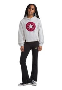 Hoodie cinza curto com um logótipo circular em bordô; combinado com calças pretas flair que apresentam um logótipo dourado. Ténis Converse em preto e branco.