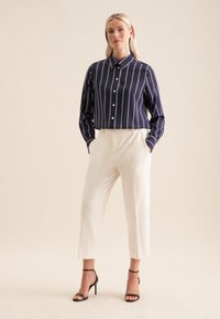 Camicia blu con bottoni e righe verticali bianche, abbinata a pantaloni a sigaretta color crema e sandali neri con tacco alto, su uno sfondo neutro.