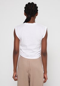 Biały, bezrękawnik z krótkim topem, okrągłym dekoltem i marszczonym tyłem, zestawiony z beżowymi, luźnymi spodniami. Gładka bawełniana tekstura.