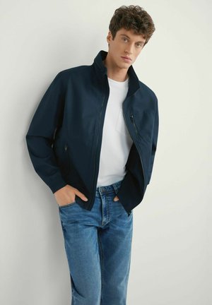 Granatowa kurtka z wysokim kołnierzem i zamkiem błyskawicznym, w połączeniu z białym t-shirtem i niebieskimi dżinsami, z bocznymi kieszeniami i gładką teksturą.