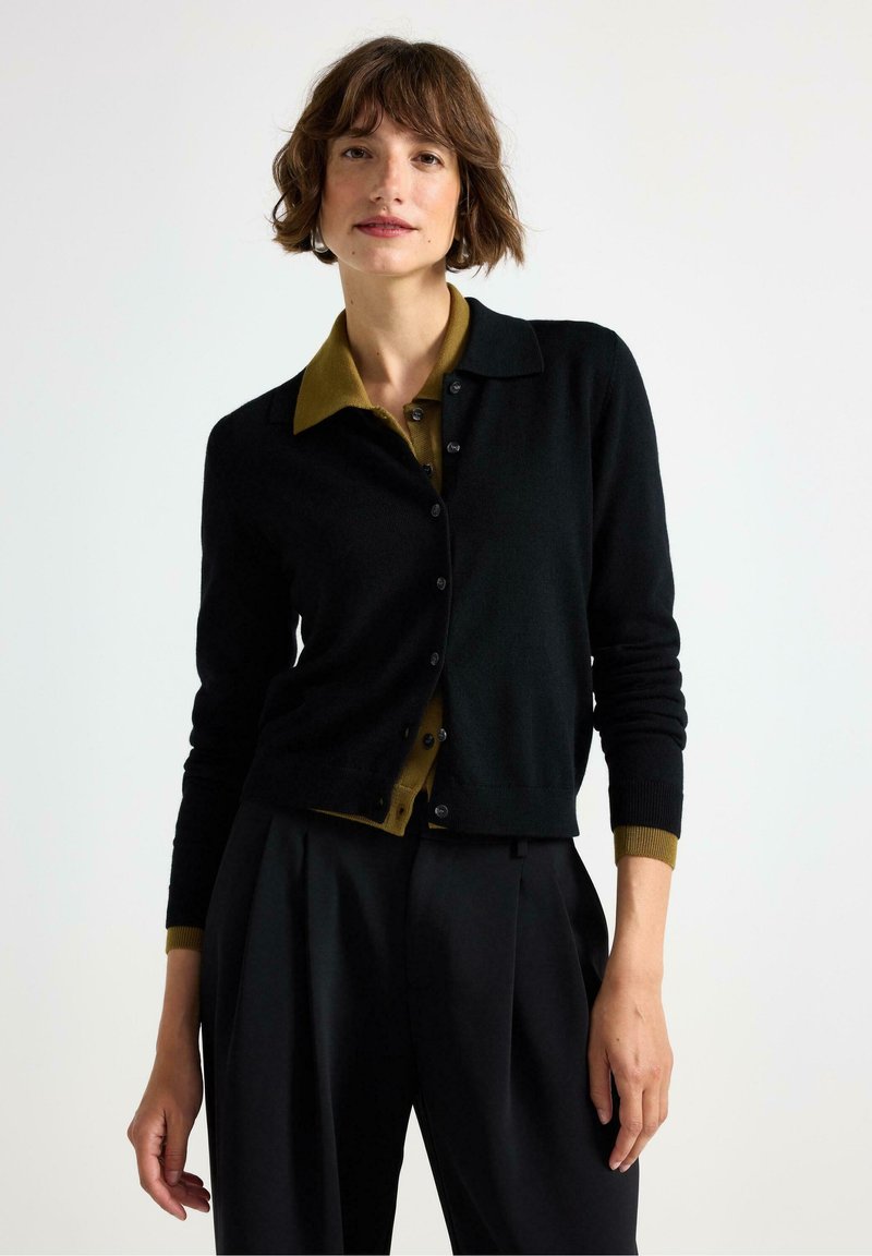 Sort cardigan med en olivengrøn skjorte med krave under, der har en knaplukningsdesign og kontrastmanchetter.