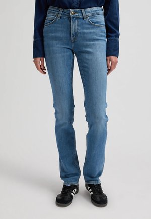 Isik, kes kannab helerohelisi kitsaid teksapükse, tumerohelist pika varrukaga pluusi ja musti tossusid valgete triipudega, seisab ühevärvilise tausta ees.