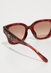 Ochelari de soare cu tentă roșie, în formă de ochi de pisică. Ramele transparente prezintă un model capitonat pe laturi și lentile gradiente de culoare maronie. Detaliu cu logo auriu.