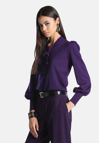 Femme aux longs cheveux foncés portant un chemisier violet avec un nœud au col, un pantalon violet assorti, une ceinture noire, de grandes boucles d'oreilles et un bracelet noir.