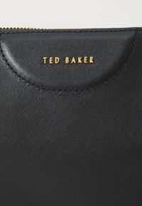 Pochette in pelle nera con logo dorato "TED BAKER", superficie testurizzata, bordo superiore arrotondato e chiusura con zip.
