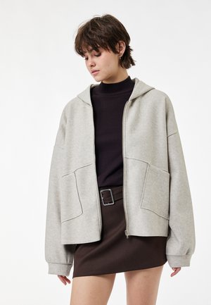 Jonge vrouw met kort haar die een lichtgrijze hoodie met rits draagt over een zwarte coltrui en een donkerbruine rok met een grote gesp riem.