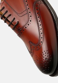 Braune Lederanzugschuhe mit Broguedetails, spitzer Zehenform, filigranen Perforationen und dunklen Schnürsenkeln auf einer glatten Oberfläche.
