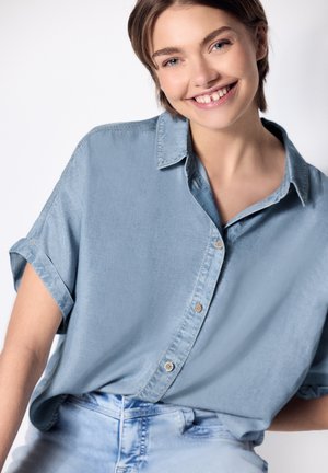 Lachende jonge vrouw die een losse lichtblauwe blouse met korte mouwen draagt en lichtgewassen spijkerbroek tegen een effen achtergrond.