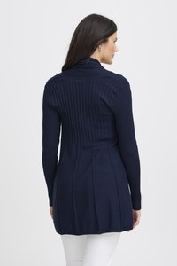 Tumesinine kudum kampsun soonikkoes, kõrge kraega ja lehviva seljaga. Pikad varrukad ja kohandatud lõige rõhutavad vormi.