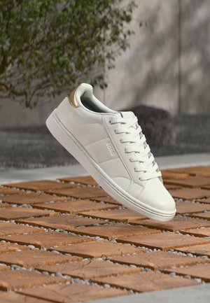 Biały, niski sneaker z złotym detalem na pięcie i sznurowadłami, prezentowany na tle drewnianej i kamiennej tekstury na zewnątrz.