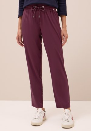 Burgundy jogger pants featuring an elastic waistband with a drawstring, smooth texture, and a loose fit, paired with white sneakers.