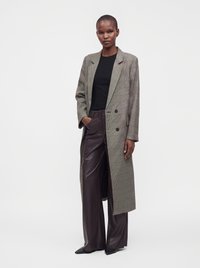 Personne debout portant un long manteau à carreaux, une chemise noire et un pantalon large en cuir, une main dans la poche, sur un fond blanc uni.