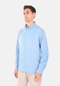 Camicia azzurra chiara, con bottoni in tessuto di cotone, colletto a punta, maniche lunghe e orlo arrotondato. Presenta bottoni bianchi lungo la parte anteriore.