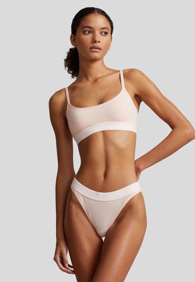 Gaiši rozā bralette un stringu komplekts, izgatavots no mīksta auduma. Iezīmējas ar plānām siksniņām un logotipa apdrukātu vidukļa joslu bez šuvēm.