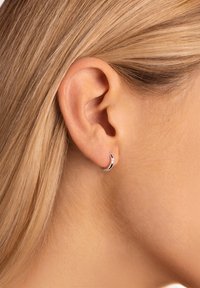 Une boucle d'oreille en argent repose sur un teint de peau clair, mettant en valeur une finition polie et une courbe subtile. Les cheveux tirés en arrière révèlent le design de la boucle d'oreille.