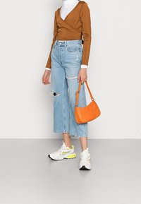 Top marrone, jeans a vita alta in denim chiaro e strappato, borsa arancione a texture e sneakers bianche con dettagli neon.