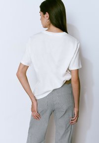 Camiseta de algodón blanca de manga corta, mostrada desde la parte trasera. Combinada con pantalones de rayas de tiro alto. Textura suave, ajuste relajado.