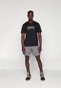 T-shirt preta de manga curta com um logótipo colorido, combinada com calções cinzentos e botas altas pretas com detalhes dourados.