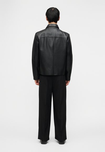 Άτομο που στέκεται με την πλάτη γυρισμένη, φορώντας μαύρο δερμάτινο μπουφάν, μαύρο παντελόνι με φαρδιά μπατζάκια και μαύρα παπούτσια, πάνω σε απλό λευκό φόντο.