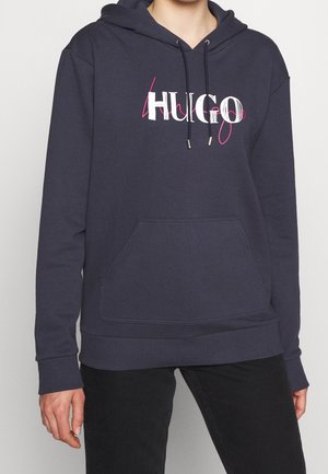 Sweat à capuche bleu marine avec poche avant, cordons de serrage et logo stylisé "HUGO" blanc et rose sur la poitrine, porté avec un pantalon noir.