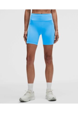Persona che indossa pantaloncini sportivi vita alta di colore blu acceso, calzini bianchi e scarpe da ginnastica bianche, in piedi davanti a uno sfondo chiaro e uniforme.