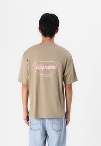 Camiseta holgada de color caqui con mangas cortas, que presenta un texto gráfico rosa en la espalda, combinada con jeans de un azul claro. Textura de algodón suave.