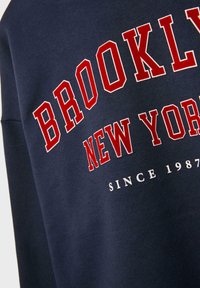 Mörkblå sweatshirt med röd och vit text "BROOKLYN NEW YORK" och mindre vit text "SINCE 1987" på framsidan.