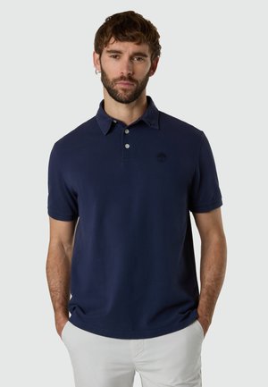 Hombre con barba que lleva una camisa polo de manga corta azul marino y pantalones de color claro, de pie con las manos en los bolsillos frente a un fondo liso.