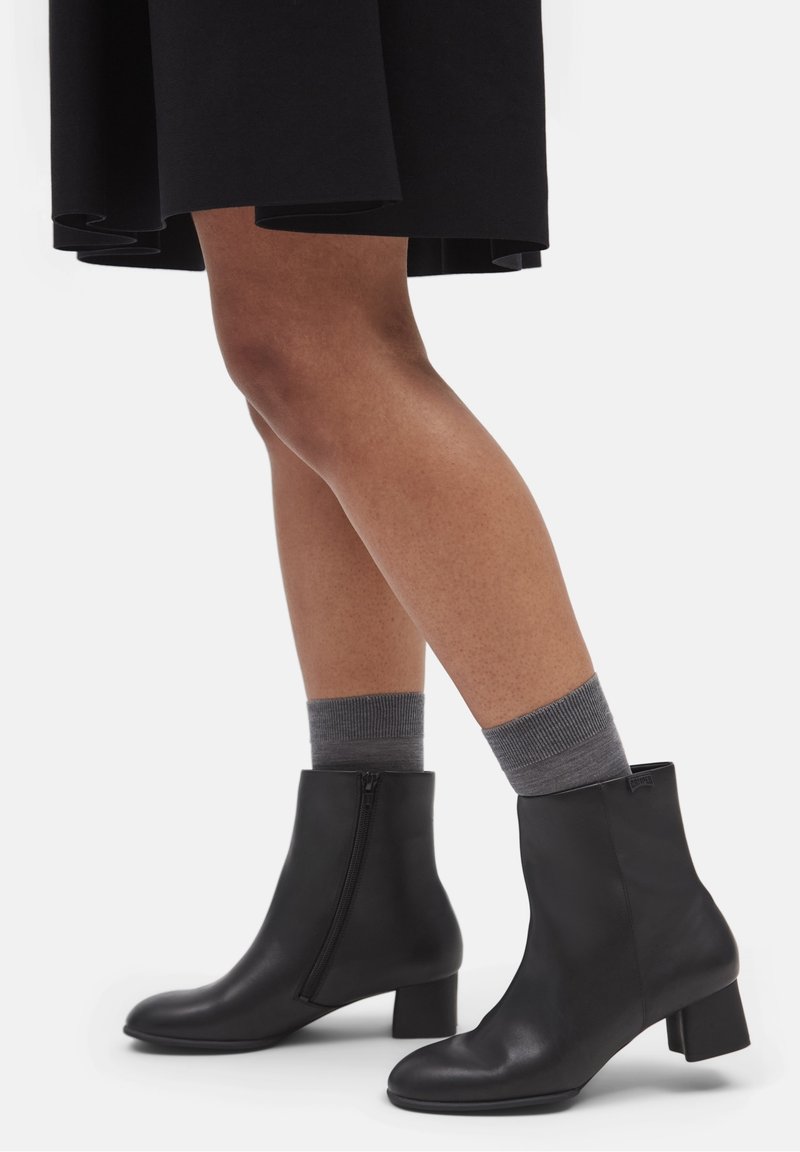 Jambes portant des chaussettes grises aux chevilles et des bottines en cuir noir à talons blocs, associées à une jupe noire mi-longue.