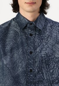 Camicia button-up in tessuto blu navy scuro con un motivo pixelato digitale, colletto strutturato e sei bottoni neri sulla parte frontale.