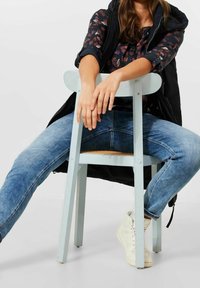 Ljust blå trä stol med vävd sits. Modell klädd i mönstrad topp och blå jeans, sitter avslappnat med händerna vilande på stolen.