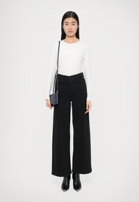 Črne široke hlače se ujemajo z oprijeto belo majico z dolgimi rokavi. Modelka drži majhno modro torbico čez ramo. Črni koničasti gležnjarji.