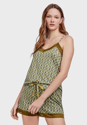 Žena nosící zelený vzorovaný komplet pyžama se spodním dílem a krátkými kalhotami s krajkovou ozdobou a páskem v pase, stojící na jednobarevném pozadí.