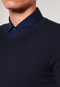 Suéter de punto azul marino con cuello redondo, patrones texturizados y una camisa con cuello de color contrastante debajo.