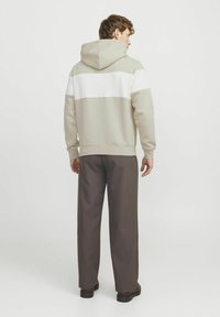 Beige hættejakke med en hvid vandret stribe, kængurulomme og snore til hætten, parret med brune bukser og sorte sko.