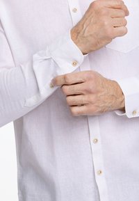 Camicia bianca di lino con design a bottoni, caratterizzata da bottoni beige e maniche arrotolate. La texture appare liscia e leggermente trasparente.